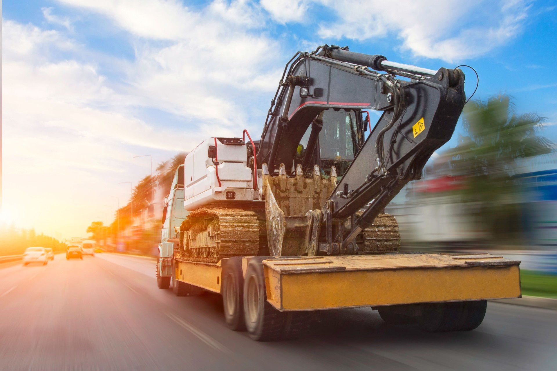 Transportation of a dirty excavator on a truck trailer after its work. Motion speed and blur effect on city highway, during sunset sky.